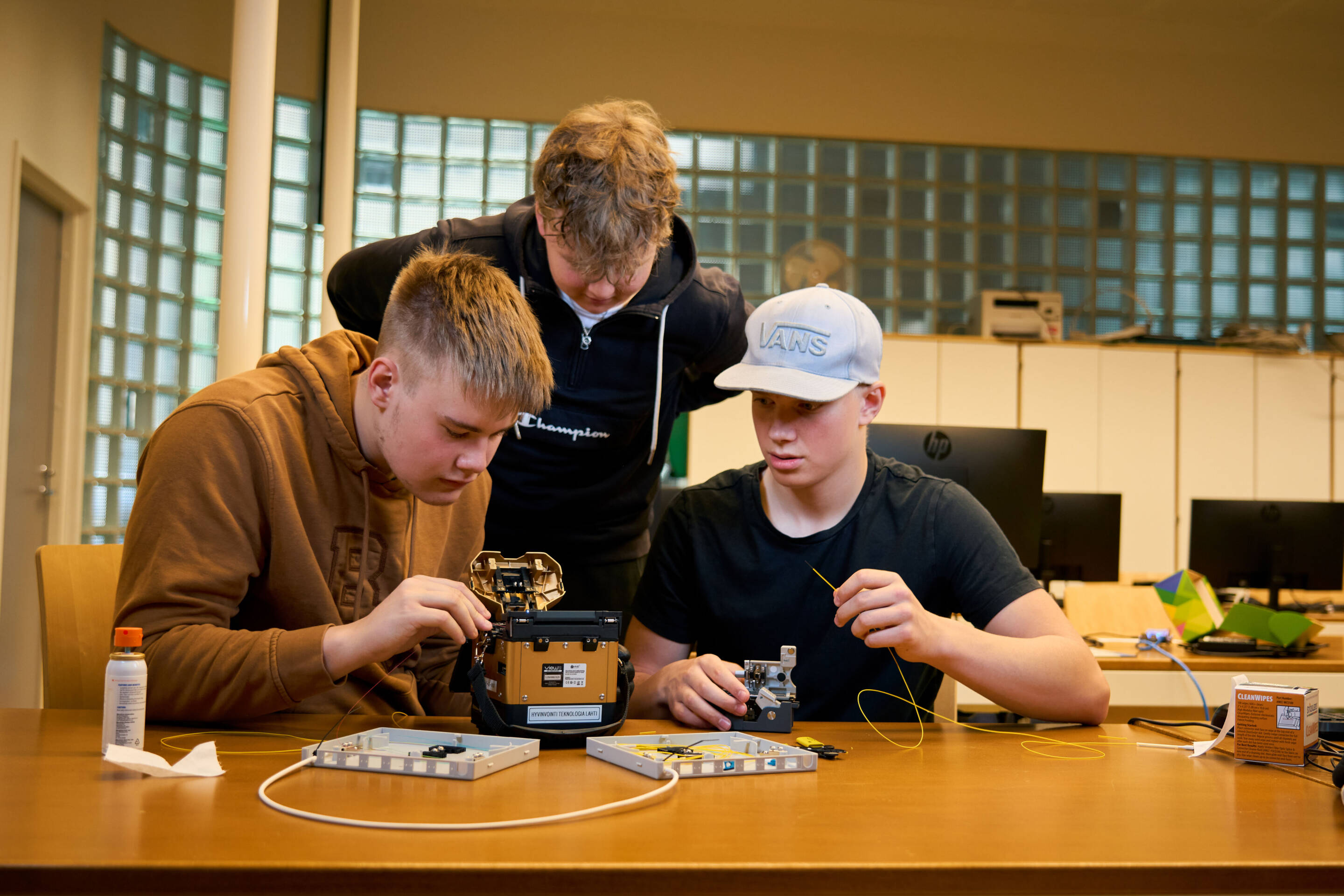 Kolme nuorta miestä osallistui tekniseen projektiin puupöydän ääressä. He käyttävät työkaluja työskennelläkseen pienellä laitteella. Toisella on huppari, toisella lippalakki ja kaikki ovat keskittyneet. Niiden takana olevista lasitiilistä tulvii tietokoneita ja valoa.