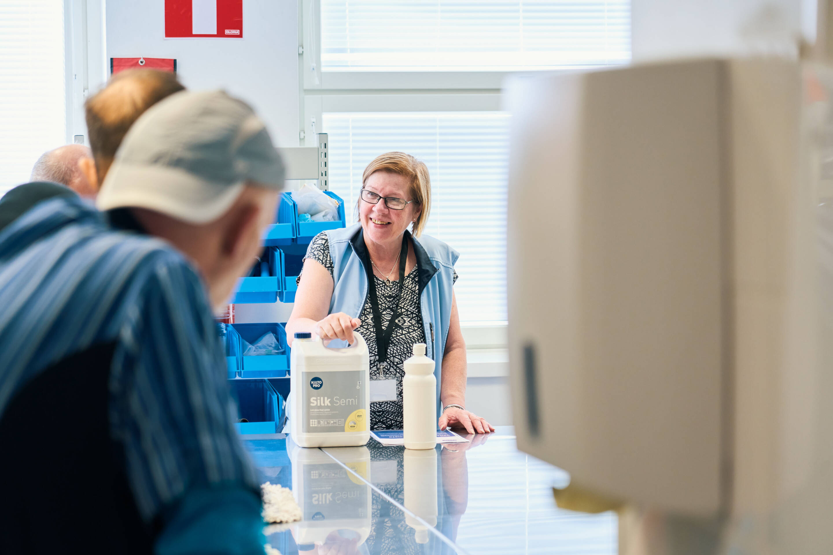 Nainen hymyilee seisoessaan työtasolla kirkkaasti valaistussa huoneessa pitäen kädessään suurta säiliötä. Muut ihmiset ovat osittain näkyvissä, osallistuvat keskusteluun. Taustalla on siniset säilytysastiat hyllyillä.