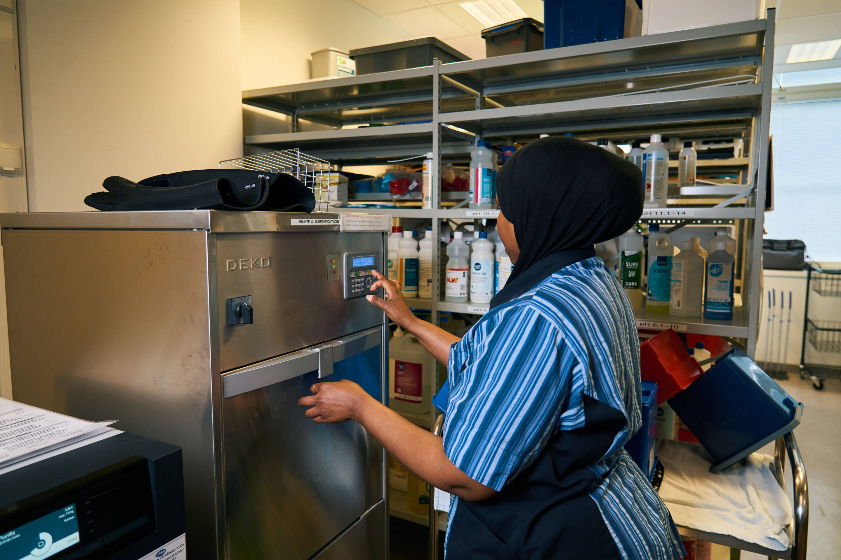 Raidallinen takki, musta huivi, käyttää ruostumattomasta teräksestä valmistettua lääketieteellistä laitetta. Niiden takana olevilla hyllyillä on erilaisia siivoustarvikkeita ja pulloja. Huone on valaistu yläpuolella olevilla loistelampuilla.