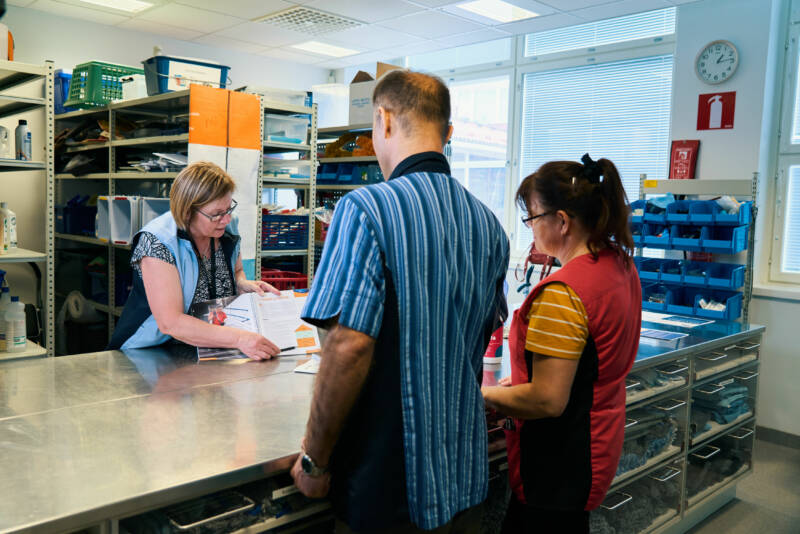 Kolme ihmistä sairaanhoitohuoneessa. Nainen tiskin takana näyttää asiakirjaa miehelle ja naiselle, jotka seisovat edessään. Taustalla näkyvät hyllyt erilaisilla tarvikkeilla ja seinällä oleva kello.