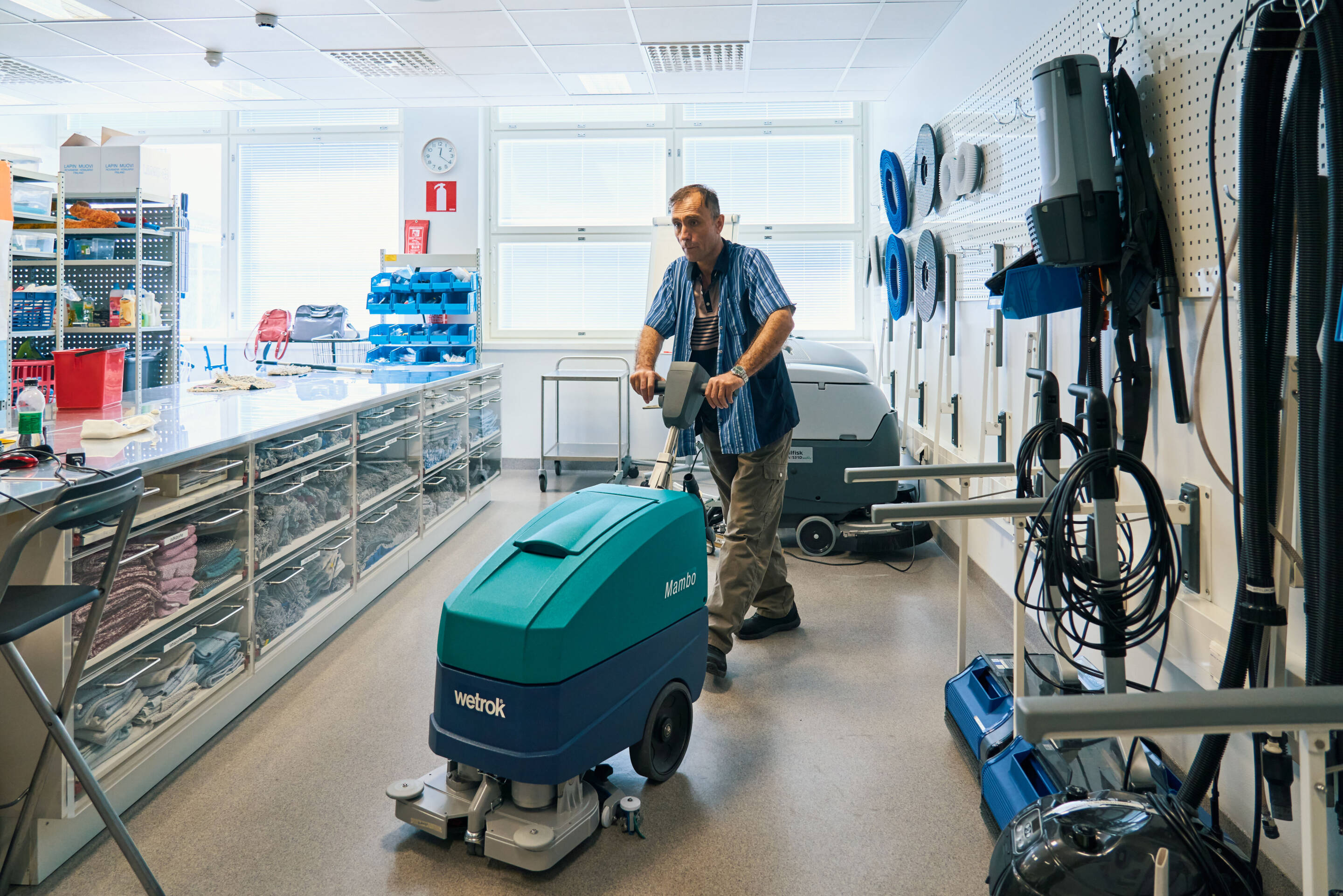 Henkilö käyttää lattianpesukonetta hyvin järjestetyssä varastotilassa, jossa on siivousvälineet ja -tarvikkeet. Huoneessa on kirkas kattovalaistus, vasemmalla hyllyt tavaroineen ja oikealla seinällä roikkuvat siivousvälineet.