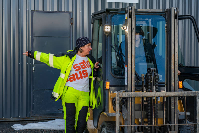 Henkilö neonkeltaisessa takissa ja housuissa, jalassa on musta huivi, elehtii vasen käsi ojennettuna seisoessaan trukin vieressä. Kohtaus sijoittuu aallotetun metallirakennuksen ulkopuolelle.
