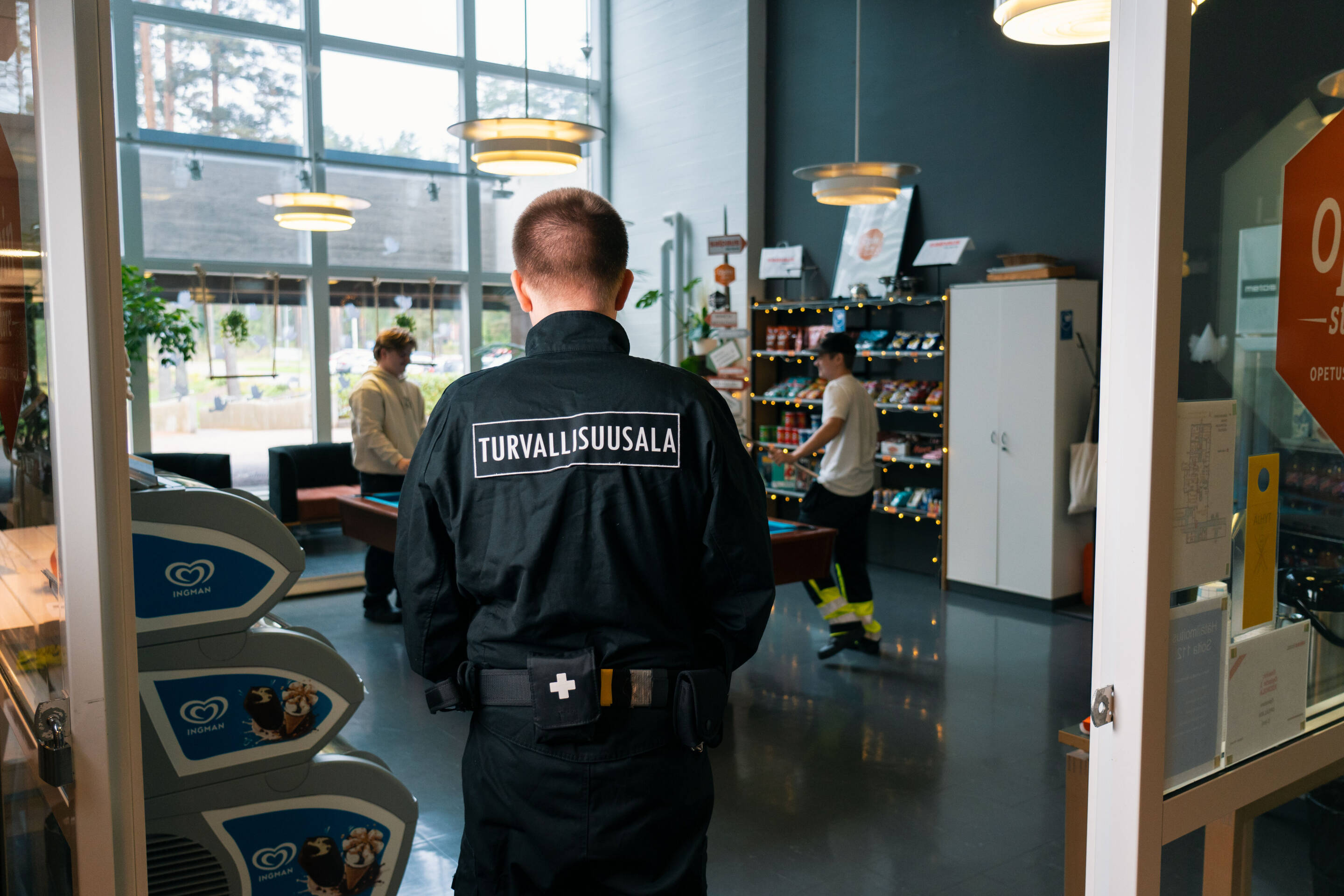 Turvallisuuspäällikkö seisoo takaa katsottuna kaupassa ja takkissaan sana Turvallisuusala. Sisällä kaksi henkilöä selailee tuotteita hyllyillä, ja luonnonvaloa tulee suurista ikkunoista.