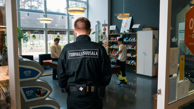 Turvallisuuspäällikkö seisoo takaa katsottuna kaupassa ja takkissaan sana Turvallisuusala. Sisällä kaksi henkilöä selailee tuotteita hyllyillä, ja luonnonvaloa tulee suurista ikkunoista.