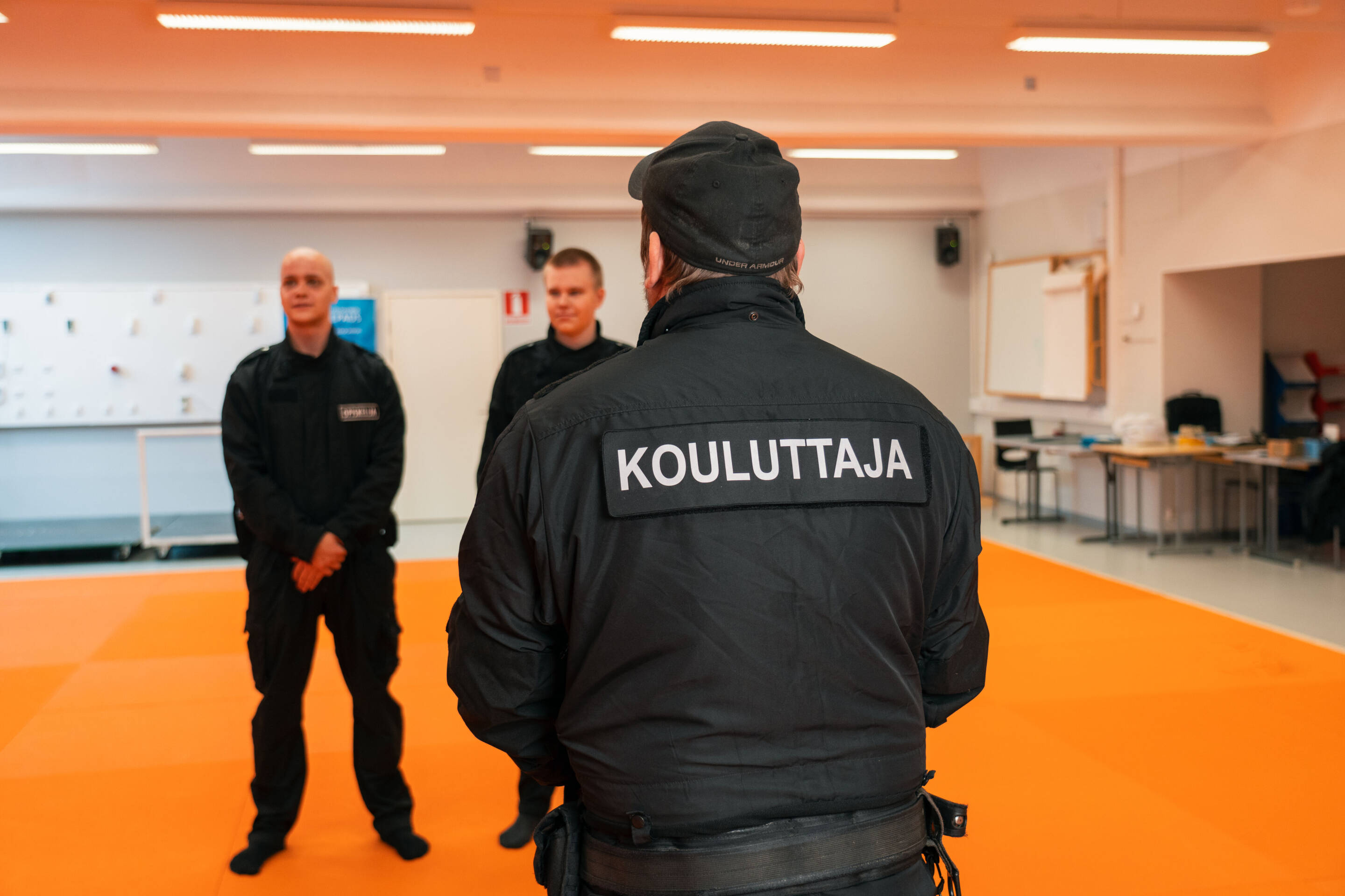 Kolme mustassa univormussa olevaa henkilöä on huoneessa, jossa on oranssi lattia. Etualalla olevan henkilön takin selkään on kirjoitettu "KOULUTTAJA", mikä osoittaa, että hän on valmentaja. Huone näyttää olevan harjoitustila.
