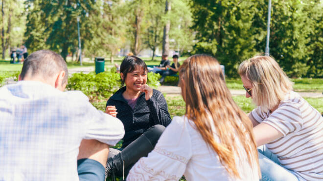 Neljän hengen ryhmä istuu nurmikolla puistossa ja nauttii toistensa seurasta. Tummatukkainen nainen hymyilee puhuessaan, kun taas muut, kuuntelevat tarkkaavaisesti. Taustalla näkyy puita ja muita ihmisiä.