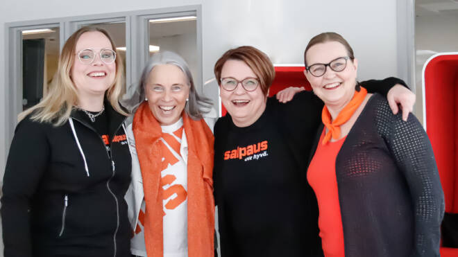 Neljä hymyilevää ihmistä seisoo tiiviisti toisissaan sisätiloissa, ja heillä on yllään arkivaatteita, joissa on Salpauksen logo. He näyttävät onnellisilta ja ovat seinän edessä, jossa on toimistoikkunat.