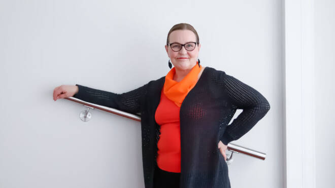 Nainen, jolla on silmälasit ja musta neuletakki oranssin topin päällä, seisoo luottavaisesti nojaten metallikaiteeseen valkoista seinää vasten. Hän hymyilee ja katsoo kameraa.