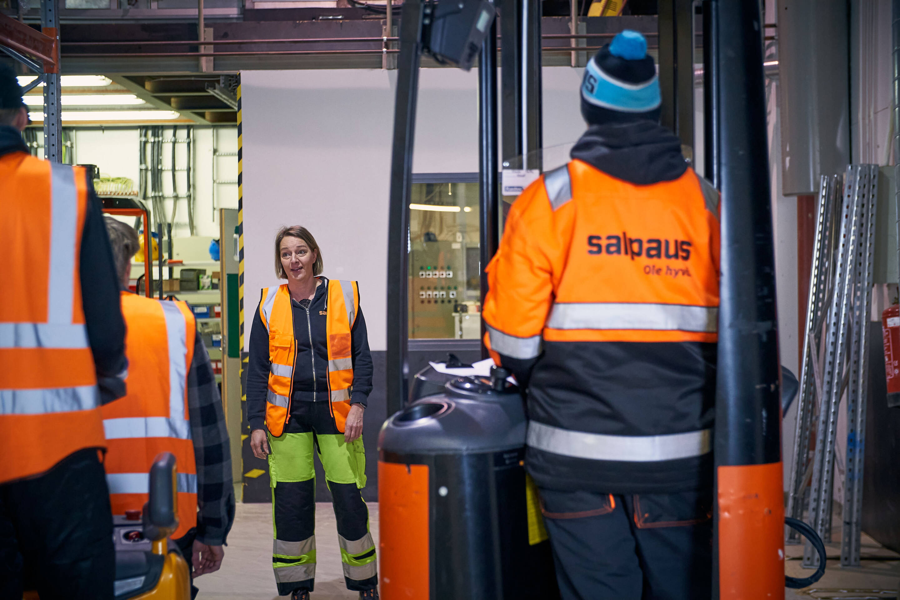 Joukko oransseissa turvaliiveissä ja lämpimissä vaatteissa pukeutuneita ihmisiä on teollisuusympäristössä. Yksi henkilö puhuu, kun taas toiset kuuntelevat trukin ympärillä. Taustalla näkyvät turvallisuus- ja teollisuuslaitteet.
