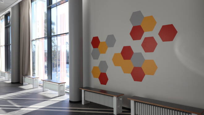 Modernin tilan sisustuksessa suuret ikkunat, valoa virtaa sisään. Seinässä on geometrinen muotoilu oransseilla, keltaisilla ja harmailla kuusikulmioilla. Seinää reunustavat metallirunkoiset puiset penkit ja lattia on harmaa laatoitettu.