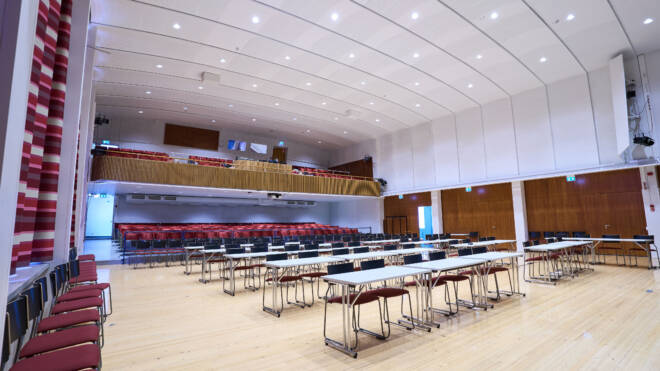 Suuri konferenssisali, jossa korkea katto, pöytä- ja tuolirivit näyttämöä päin. Auditorioalueen täyttävät punaiset tuolit ja näkyvissä on parveke lisäistuimilla. Huoneessa on moderni valaistus ja puulattia.