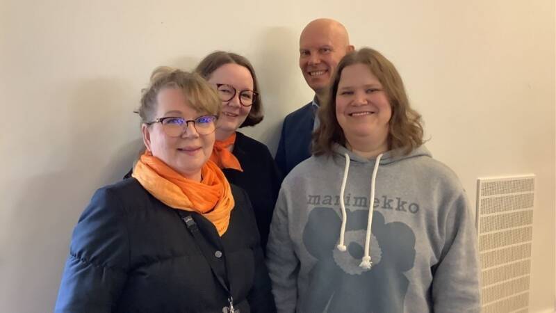 Neljä aikuista seisoo hymyillen vaaleanvärisen seinän edessä. Edessä on kolme naista, joista kahdella on oranssit huivit ja yhdellä harmaa huppari. Heidän takanaan seisoo hymyilevä mies.