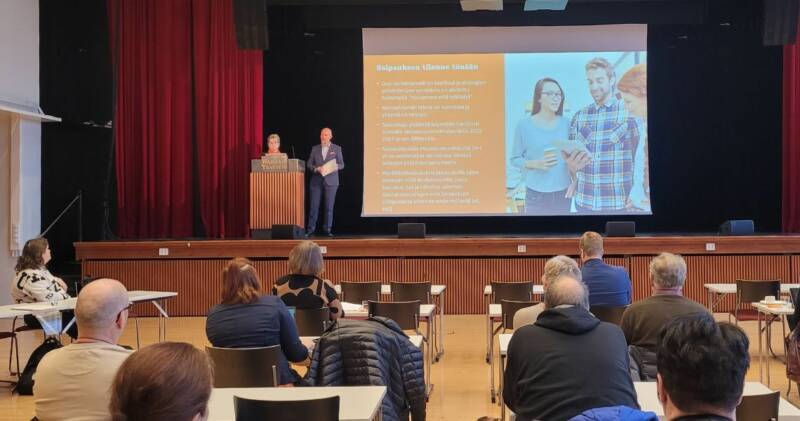 Esitys pidetään salissa, jossa ihmiset istuvat lavaa päin. Kaksi puhujaa seisoo korokkeella suuren näytön vieressä, jossa näkyy tekstiä ja kuvia kolmesta ihmisestä. Yleisö kuuntelee tarkkaavaisesti.