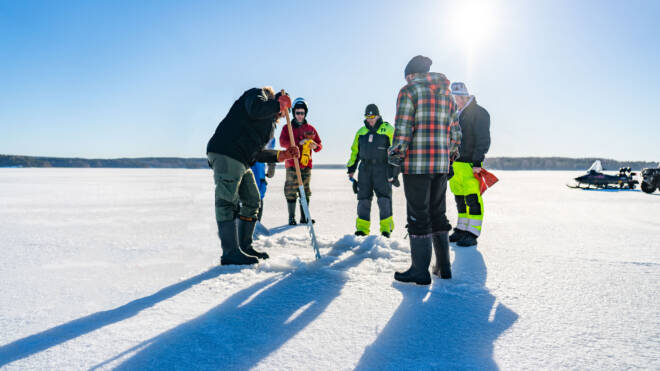 Ryhmä talvivaatteita pukeutuneita ihmisiä seisoo jäätyneellä järvellä. Yksi henkilö poraa jään sisään kairan avulla. Kirkas auringonvalo luo pitkiä varjoja ja taustalla näkyy kelkka. Taivas on kirkas ja sininen.