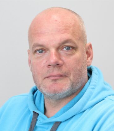 Keski-ikäinen mies, jolla on ajeltu pää ja lyhyt harmaa parta, on pukeutunut vaaleansiniseen huppariin. Hän katsoo suoraan kameraan neutraalilla ilmeellä yksinkertaista vaaleaa taustaa vasten.