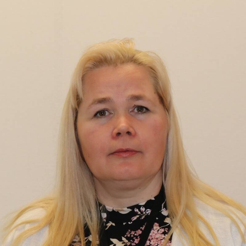 Pitkät vaaleat hiukset omaava nainen, jolla on musta kukkakuvioinen toppi ja valkoinen neule, katsoo suoraan kameraan yksinkertaista, vaaleaa taustaa vasten.