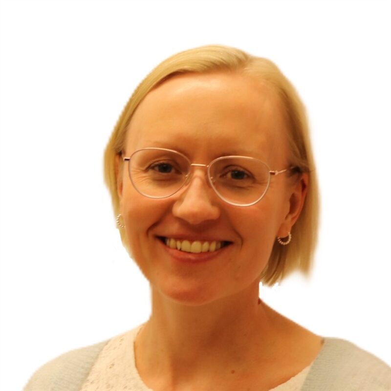 Henkilö, jolla on lyhyet vaaleat hiukset ja silmälasit, hymyilee. Heillä on yllään vaaleansininen neuletakki valkoisen topin päällä. Tausta on tavallinen valkoinen.
