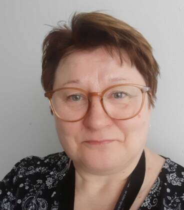 Lyhythiuksinen ja silmälasipäinen henkilö hymyilee hieman kameralle. Hänellä on yllään mustavalkoinen kuviollinen paita ja musta kaulanauha, ja hän seisoo vaaleanvärisen seinän edessä.