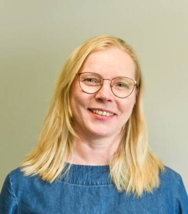 Nainen, jolla on pitkät vaaleat hiukset ja silmälasit, hymyilee kameralle. Hänellä on yllään sininen toppi ja hän seisoo tavallista vaaleaa taustaa vasten.