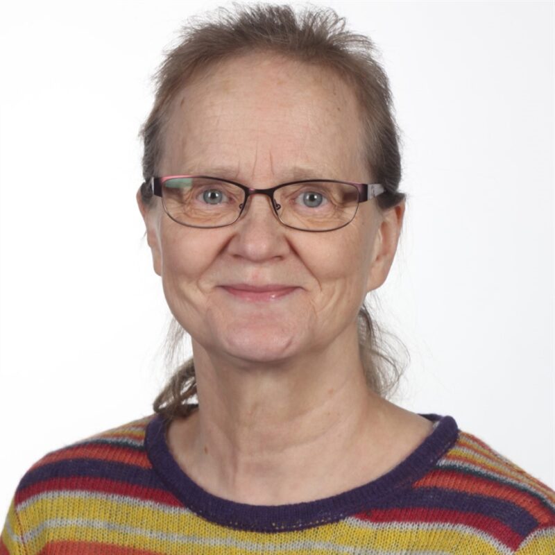 Hymyilevä, vaaleaihoinen, silmälasit käyttävä ja taaksepäin vedetyt ruskeat hiukset omaava vanhempi nainen poseeraa tavallisen valkoisen taustan edessä. Hänellä on yllään värikäs raidallinen neulepusero.