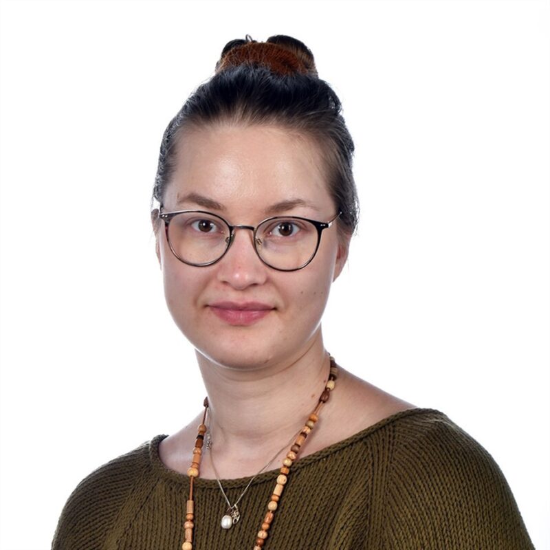 Henkilö, jolla on ruskeat hiukset nuttulassa, silmälasit ja vihreä pusero. Heillä on kaulakoru, ja ne poseerataan tavallista valkoista taustaa vasten.