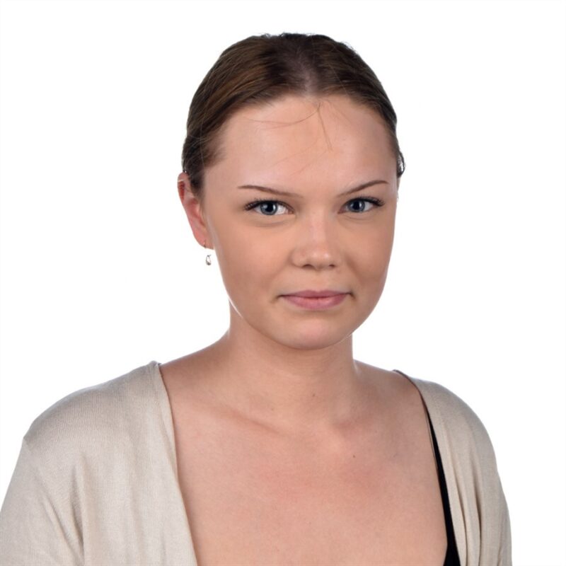 Vaaleanruskeat hiukset taakse vedetyllä henkilöllä on yllään beige toppi mustan paidan päällä. Heillä on neutraali ilme ja he katsovat suoraan kameraan tavallista valkoista taustaa vasten.