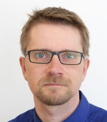 Mies, jolla on lyhyet, vaaleanruskeat hiukset ja pukinhakki, käyttää mustakehystettyjä laseja ja sinikauluspaitaa ja poseeraa valkoista taustaa vasten.