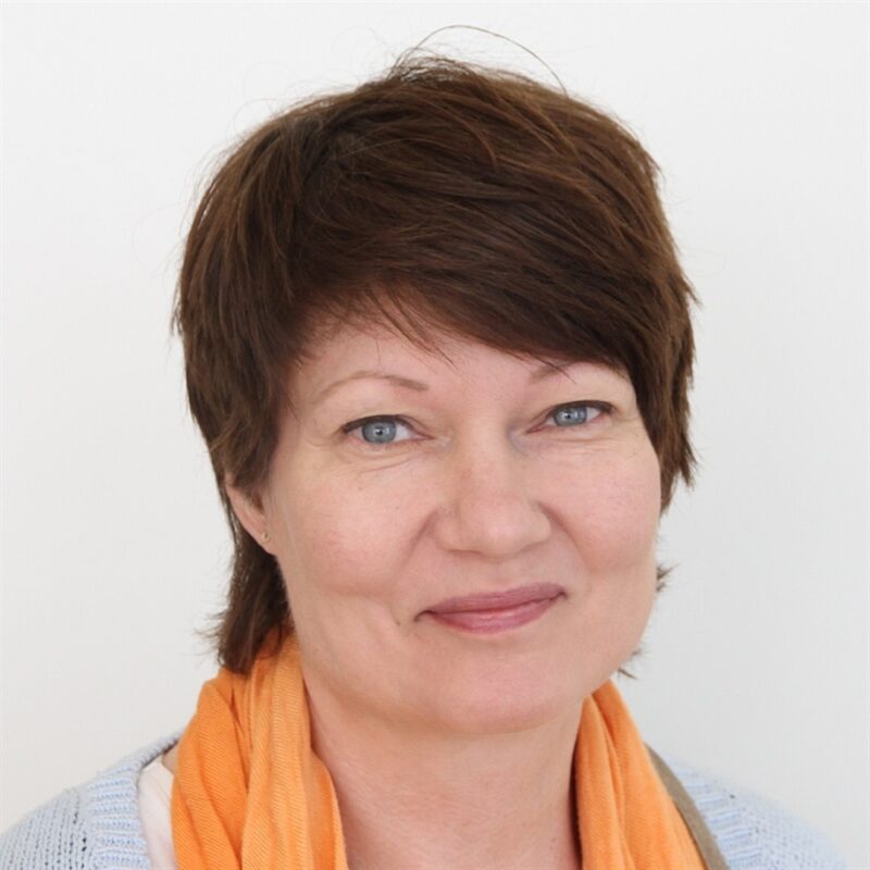 Henkilö, jolla on lyhyet ruskeat hiukset, hymyilee hieman valkoista taustaa vasten. Heillä on yllään vaaleanharmaa toppi ja oranssi huivi.