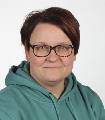 Henkilö, jolla on silmälasit ja sinivihreä huppari, hymyilee kameralle tavallista valkoista taustaa vasten.