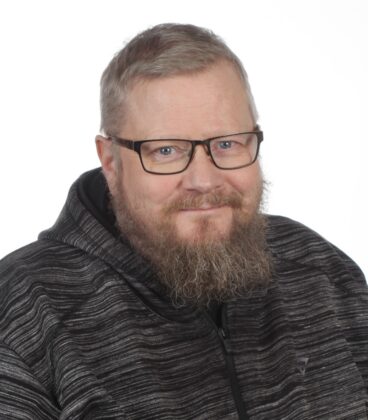 Parrakas mies, jolla on silmälasit ja tumma, raidallinen huppari, hymyilee hieman katsoessaan kameraa yksinkertaista valkoista taustaa vasten.