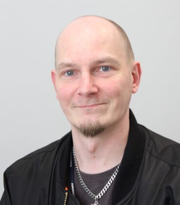 Henkilöllä, jolla on ajeltu pää ja vuohenpaita, on yllään musta takki ja hopeaketjukaulakoru, ja hän hymyilee kevyesti valkoista taustaa vasten.