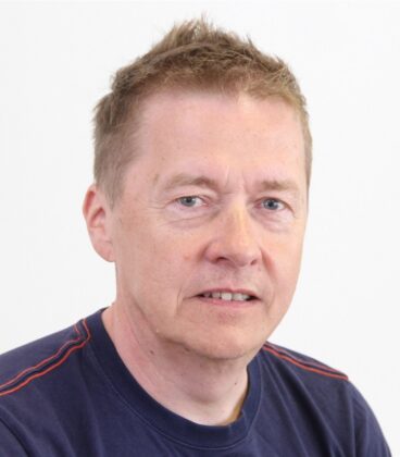 Keski-ikäinen mies, jolla on lyhyet, vaaleanruskeat hiukset ja tummansininen paita, istuu valkoisen taustan edessä ja katsoo kameraa neutraalilla ilmeellä.