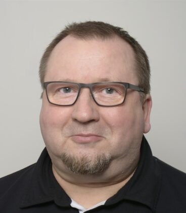 Keski-ikäinen mies, jolla on lyhyet vaaleanruskeat hiukset, silmälasit ja vaalea vuohenparkki hymyilee hieman kameralle. Hänellä on yllään musta paita ja hänellä on neutraali harmaa tausta.