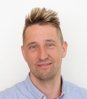 Henkilö, jolla on lyhyet, piikkihiukset ja ohut parta, hymyilee kameralle. Heillä on yllään vaaleansininen napillinen paita. Tausta on tavallinen valkoinen.