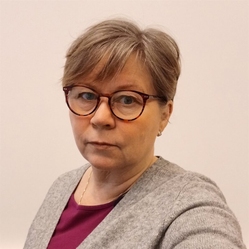 Henkilö, jolla on lyhyet, harmaat hiukset ja silmälasit, jolla on harmaa pusero violetin topin päällä. Tausta on tasainen ja vaalea.