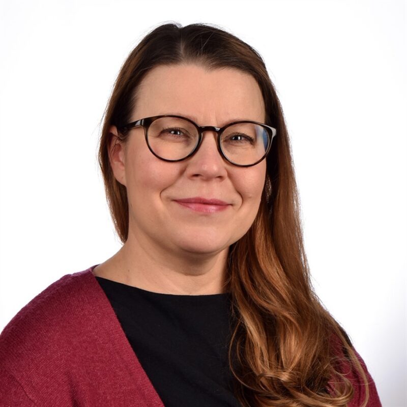 Nainen, jolla on pitkät, aaltoilevat ruskeat hiukset, silmälasit, musta toppi ja kastanjanruskea neuletakki, hymyilee pehmeästi. Hänet on kuvattu valkoista taustaa vasten.