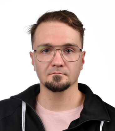 Mies, jolla on lyhyet ruskeat hiukset, parta ja silmälasit, yllään musta takki ja vaaleanpunainen paita, seisoo valkoista taustaa vasten.
