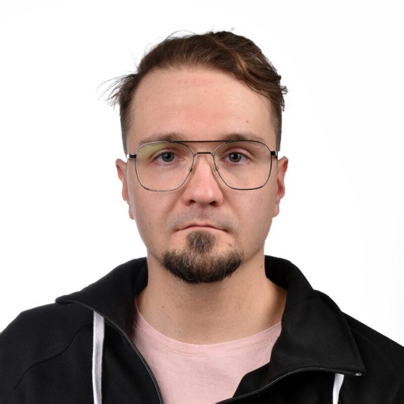 Mies, jolla on lyhyet ruskeat hiukset, parta ja silmälasit, yllään musta takki ja vaaleanpunainen paita, seisoo valkoista taustaa vasten.