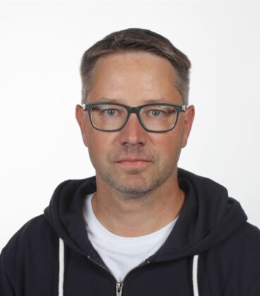 Lyhyethiuksinen henkilö, jolla on silmälasit ja tumma huppari valkoisen paidan päällä, poseeraa muotokuvassa tasaista taustaa vasten.