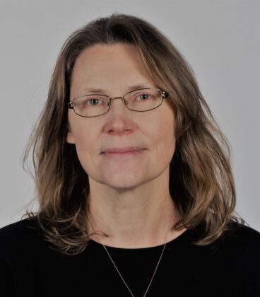 Henkilö, jolla on olkapäille ulottuvat hiukset ja silmälasit, yllään musta toppi ja kaulakoru, poseeraa tavallista taustaa vasten.