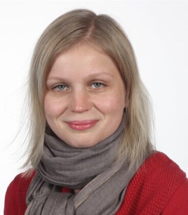 Nainen, jolla on olkapäille ulottuvat vaaleat hiukset, hymyilee kameralle. Hänellä on yllään punainen toppi ja harmaa huivi. Tausta on tavallinen valkoinen.