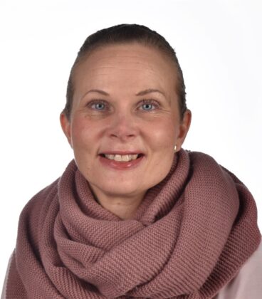 Vaaleaihoinen ja sinisiilmäinen nainen hymyilee kameralle. Hänellä on yllään paksu, neulottu malvanvärinen huivi ja vaalea toppi. Hänen hiuksensa on vedetty taaksepäin ja tausta on yksivärinen valkoinen.
