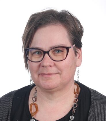 Lyhythiuksinen ruskeatukkainen nainen, jolla on silmälasit, tumma paita, harmaa neuletakki ja paksu kaulakoru, hymyilee hieman kameralle yksinkertaista valkoista taustaa vasten.