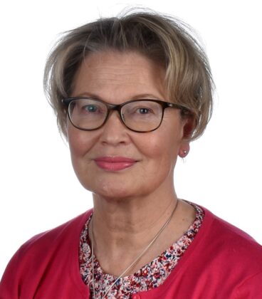 Henkilö, jolla on lyhyet, vaaleanruskeat hiukset ja silmälasit, hymyilee. Heillä on yllään punainen takki kukkapuseron päällä. Tausta on tavallinen valkoinen.