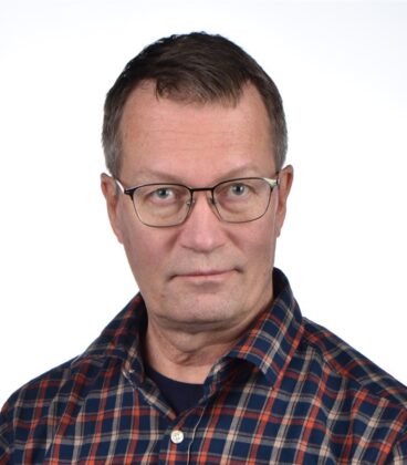 Silmälasit ja ruudullinen paita pukeutunut henkilö katsoo suoraan kameraan valkoista taustaa vasten.