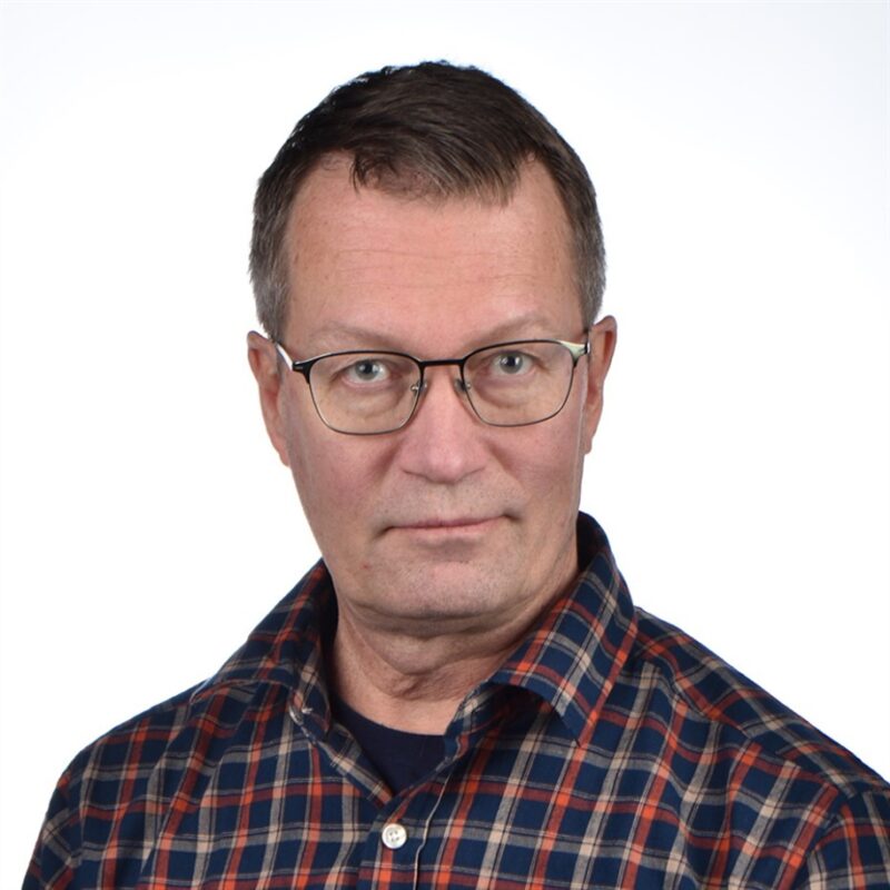 Silmälasit ja ruudullinen paita pukeutunut henkilö katsoo suoraan kameraan valkoista taustaa vasten.