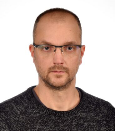 Mies, jolla on silmälasit ja parta, yllään tumma villapaita, katsoo suoraan kameraan. Tausta on tavallinen valkoinen.
