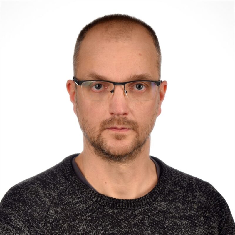 Mies, jolla on silmälasit ja parta, yllään tumma villapaita, katsoo suoraan kameraan. Tausta on tavallinen valkoinen.