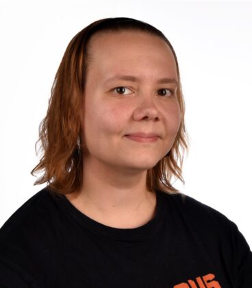 Hartioihin ulottuvat vaaleanruskeat hiukset hymyilevät hieman, ja hänellä on musta paita valkoista taustaa vasten.