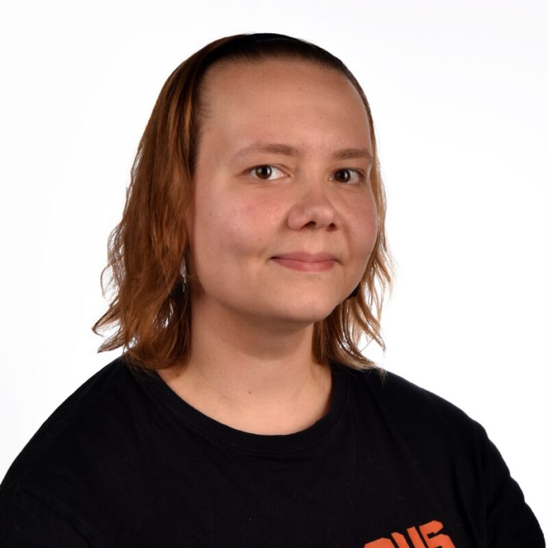 Hartioihin ulottuvat vaaleanruskeat hiukset hymyilevät hieman, ja hänellä on musta paita valkoista taustaa vasten.